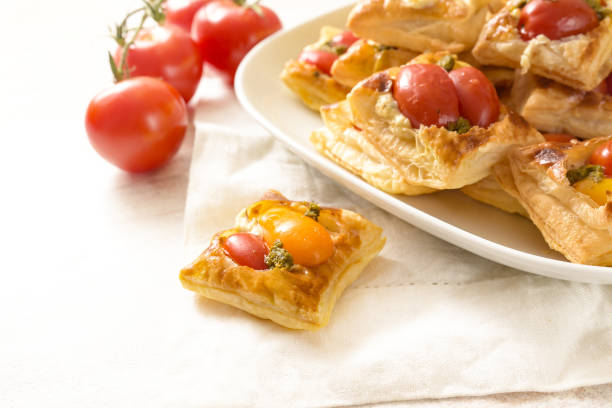 Baked puff pastry snacks with cream cheese, tomatoes and herb pesto, party finger food served on a white napkin and a plate at a cold buffet, copy space
