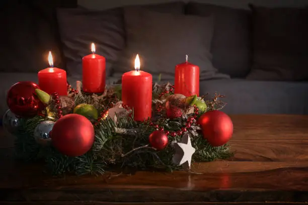 Advent wreath with three burning red candles and Christmas decoration on a wooden table in front of the couch, festive home decor for the third Sunday, copy space, selected focus, narrow depth of field