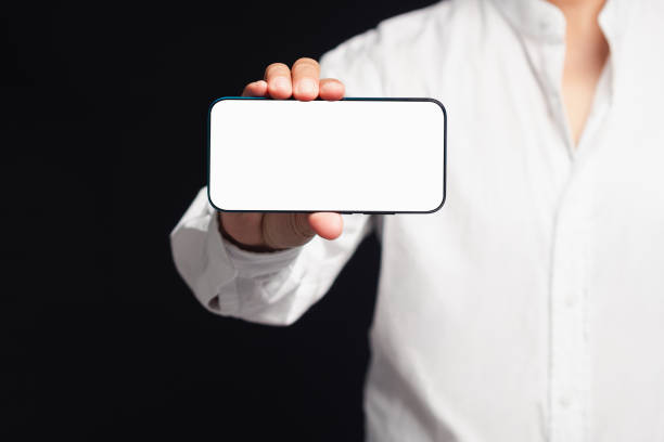 istock Businessman holding a horizontal mobile phone with a blank white screen while standing on a black background 1740528728