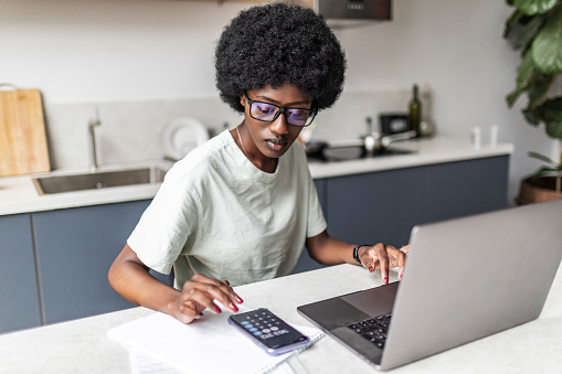 Young adult black woman working from home at the kitchen using laptop and mobile