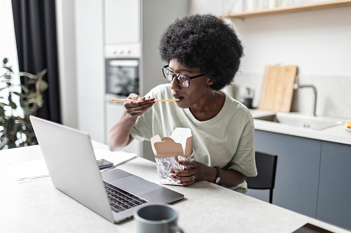 Young adult black woman working from home using laptop and eating at the same time take out asian food