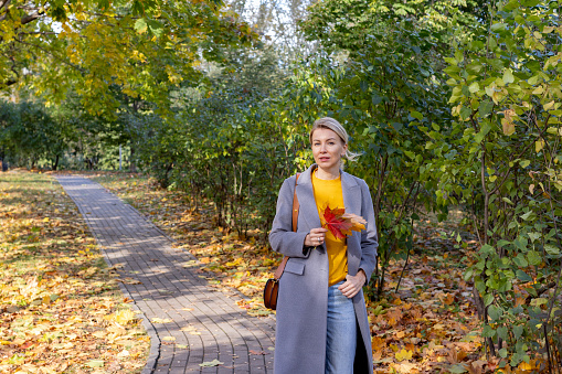 Middle age trendy woman in gray coat and jeans walking in autumn park