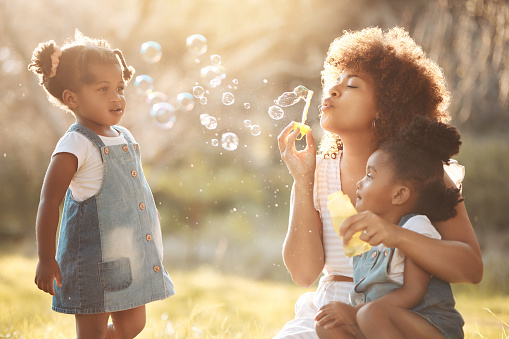Nature, children and mother blowing bubbles in an outdoor park for playing, bonding or having fun. Happy, love and young mom sitting with her girl kids in a field or green garden in summer in Mexico.