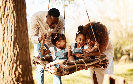 Parents with kids in park, tree swing and freedom for love, bonding or peace in nature together. Mother, father and children adventure in garden, black family on summer weekend in woods or forrest.