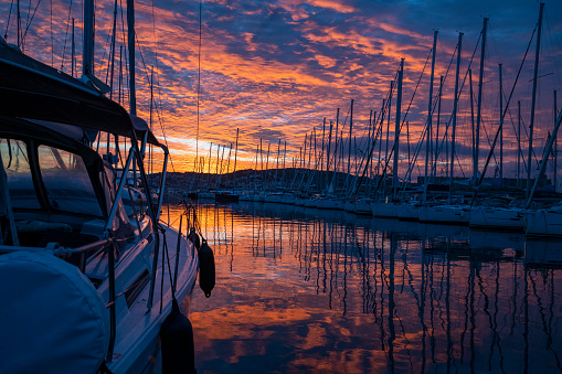 Sailing ship in port at sunset with red sky and sailing boats reflection on water.