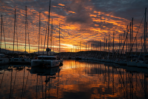 Sailing ships in port at sunset with red sky and sailing boats reflection on water.
