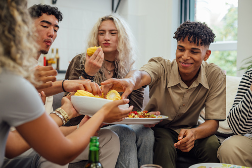 An over the shoulder view of a group of young university students sitting in the common room of their university enjoying some Mexican food together and enjoying some non alcoholic beers together. A member of the group who is non binary is enjoying some fresh cooked corn.