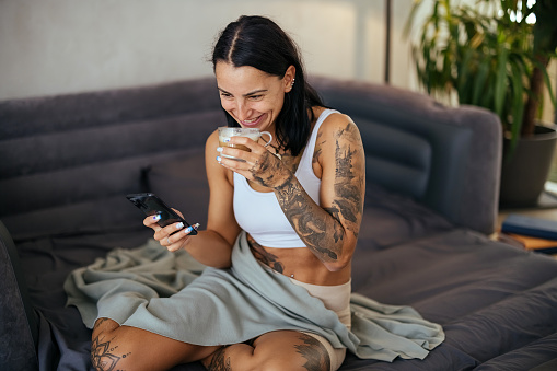 Young woman drinks coffee and uses a mobile phone while sitting on the bed in the morning