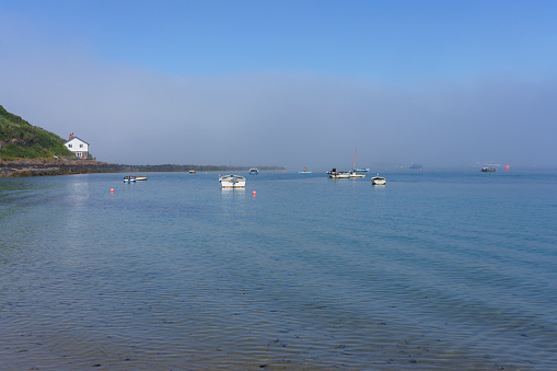 Small boats are moored in Porthdinllaen harbour under a blue sky while out a sea a thick bank of fog lingers.
