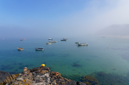 Fog swirls around boats moored on the crystal clear water of Porthdinllaen harbour lit by bright summer sunshine.