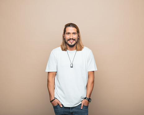 Happy mid adult man wearing white t-shirt standing with hands in pockets and smiling at camera. Studio shot, beige background.