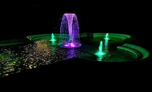 the fountain sprays strips of water intertwined into one column. the colors are bright pastels. historic swimming pool at the castle garden at night, water jet, illumination