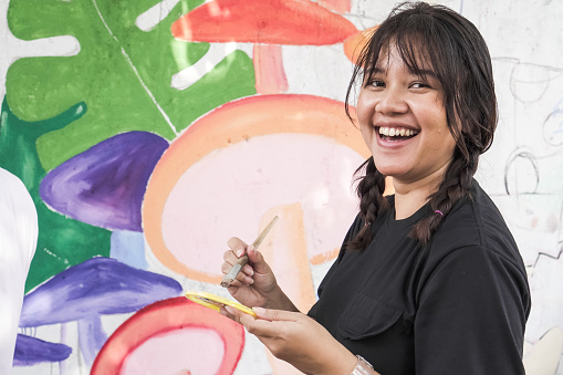 Yogyakarta, Indonesia - August 8, 2023: Happy beautiful asian female artist is drawing plants mushrooms and flowers mural on the white wall with brush. 17 Agustus Decoration, Indonesian Independence Day Concept