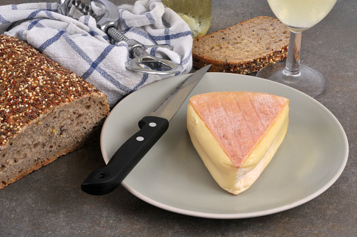 Piece of Saint Nectaire cheese on a plate with a knife and seeded bread and a glass of wine
