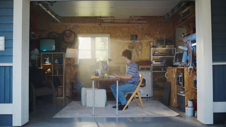 Caucasian Male Software Engineer Programming On Old Desktop Computer In Retro Garage With Random Appliances. Man Working On Innovative Online Service Startup Company In Nineties. Nostalgia Concept.