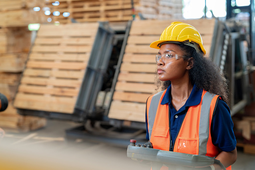 African American technician wear protective workwear, stands at the forefront of the production line, using a remote control to orchestrate the precise movements of robotic arm. Fusion of human expertise with technology is on full display. Ensuring quality control and the continual improvement of processes.