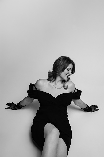 A female model in a black dress poses in a photo studio on a white background. Monochrome shot.