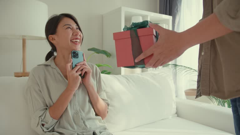 Happy Asian couple having fun surprise gifting birthday present in red gift box sit on sofa in living room at home. Lifestyle spend time in house.