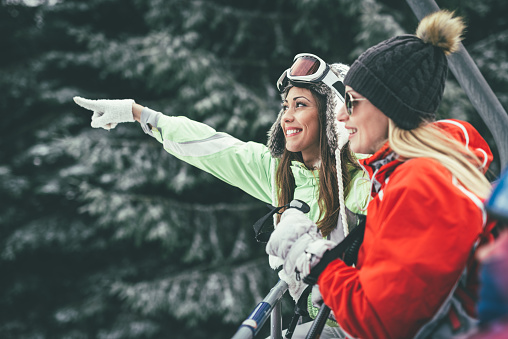 Beautiful young woman friends enjoying in winter vacations. They driving on ski lift and looking away with smile.