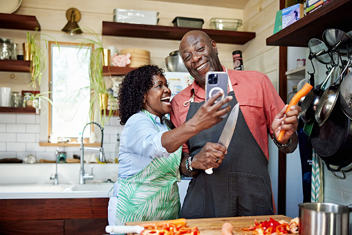 Waist-up view of senior Black man holding kitchen knife and carrot, mature woman holding smart phone, both in aprons and laughing.