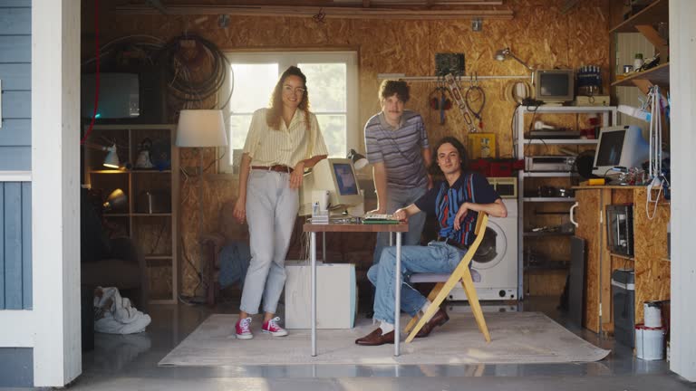 Diverse Team Of Three Young Startup Company Founders In Retro Garage With Old Desktop Computer. Male And Female Entrepreneurs Looking At Camera And Smiling. Developing Online Service In Nineties.