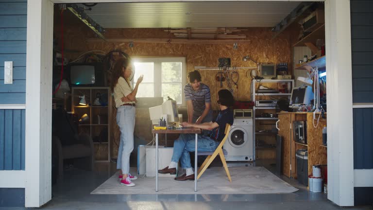 Diverse Team Of Three Young Startup Company Founders Working In Retro Garage, Using Old Desktop Computer. Male And Female Entrepreneurs Developing Innovative Online Service In Nostalgic Nineties.