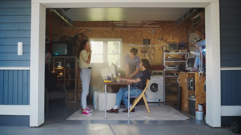 Diverse Team Of Three Young Tech Startup Company Founders Working In Retro Garage, Using Old Desktop Computer. Male And Female Entrepreneurs Developing Innovative Online Service In Nostalgic Nineties.