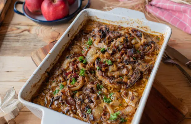 Delicious autumn winter food recipe with oven bake pork chops in a tasty onion, bacon cream sauce. Served hot and ready to eat in a baking dish on rustic and wooden table background. Closeup