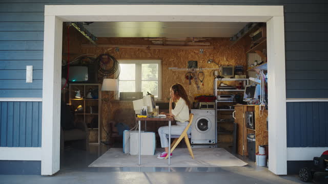Hispanic Female Developer Programming On Old Desktop Computer In Retro Garage With Random Appliances. Young Woman Working On Innovative Online Service Startup Company In Nineties. Nostalgia Concept.