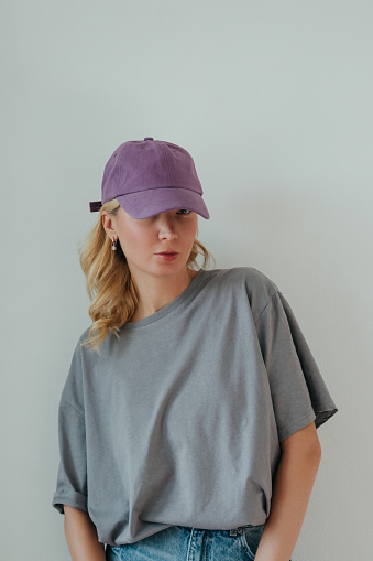 Young woman dressed oversized T-shirt, jeans, cap, stands against wall, looking the camera.