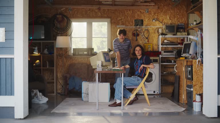 Two Caucasian Male Startup Founders Using Old Desktop Computer In Retro Garage, Looking At Camera And Smiling. Programmer And UX Designer Starting Internet Business In Nineties. Nostalgia Concept