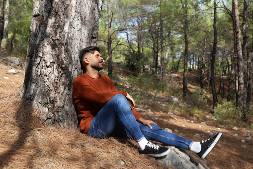 Bearded guy sitting in the woods alone and relaxing. Young man sitting under the tree in meditative state, leaning on the trunk. Close up, copy space, forest landscape background.