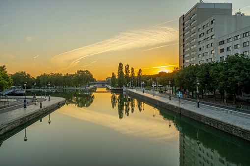 Paris, France - 08 15 2021: Ourcq Canal. Reflections on the Ourcq canal of a bridge, trees, barges and buildings at sunrise