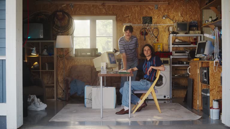 Two Caucasian Male Tech Startup Founders Using Old Desktop Computer In Retro Garage, Looking At Camera And Smiling. Programmer And UX Designer Starting Internet Business In Nineties. Nostalgia Concept