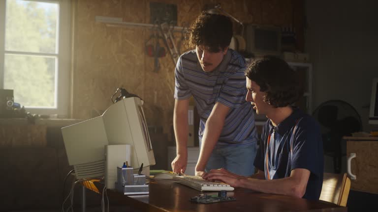 Two Caucasian Male Tech Startup Founders Using Old Desktop Computer In Retro Garage. Software Developer And User Experience Designer Starting New E-commerce Business In nineties. Nostalgia Concept.