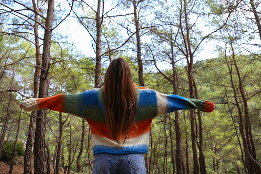Back view of a young woman in a hippie style outfit standing with spread arms, absorbing sun energy in the woods. Close up, copy space, forest landscape background.