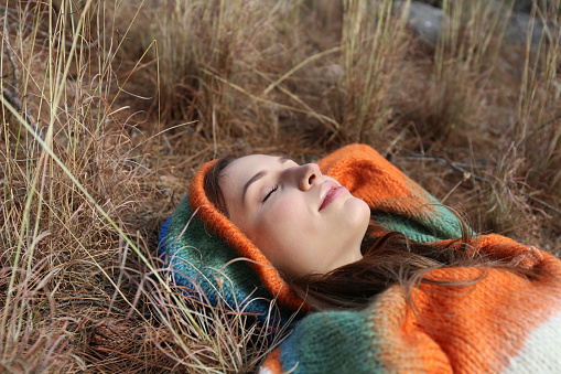 Portrait of a relaxed young woman wearing a colorful knitted hoodie lying on the ground in the woods. Close up, copy space, background.