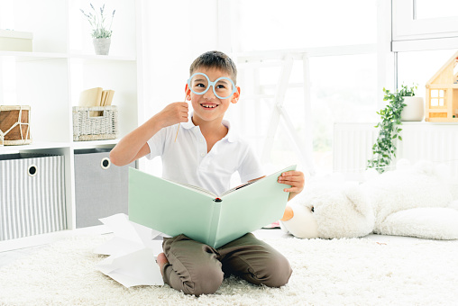 Happy little boy reading book with glasses on stick in children's room