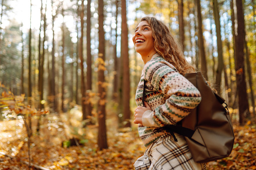 Beautiful young tourist with a backpack is having fun in the autumn forest, throwing fallen leaves. Woman walks along a colorful path in the forest. Freedom, travel.
