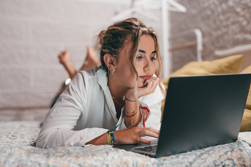 Close up of one young pretty woman using laptop indoor at home lying on the bed surfing the net. Millennial teenager having fun online studying.