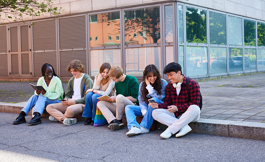 Diverse group of teenager students using smart phones and reading books while sitting on footpath in college campus