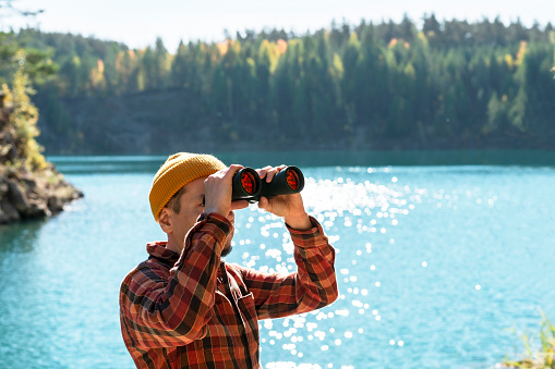 young man, dressed in red plaid shirt, engaged in birdwatching with binoculars on the banks of calm blue mountain lake in the autumn forest A haven for nature enthusiasts copy space
