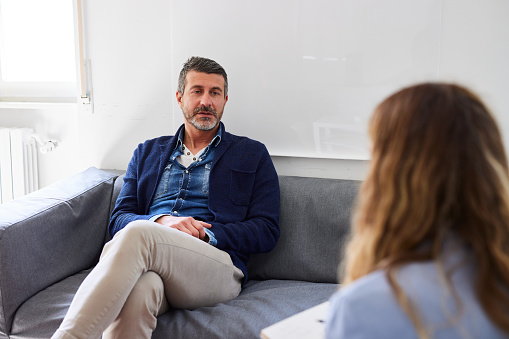 Mid adult man sharing his marriage problems with female psychologist during therapy session while sitting on sofa in doctor's office