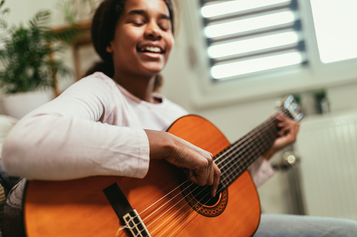 African american teenage girl sitting on couch in her room and learning to play guitar