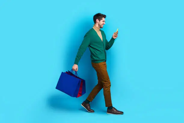 Full body photo of young working man brunet hair walking going with phone pre order adidas new collection isolated on blue color background.