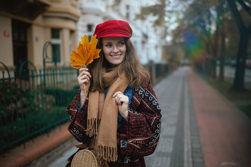 Hello autumn. smiling modern woman in red hat with autumn leafs and scarf walking in the city.