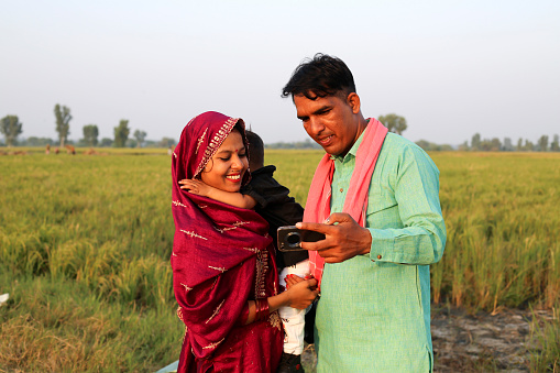 Cheerful Indian farmer family standing portrait near green field during springtime also using mobile phone and taking selfie using mobile camera