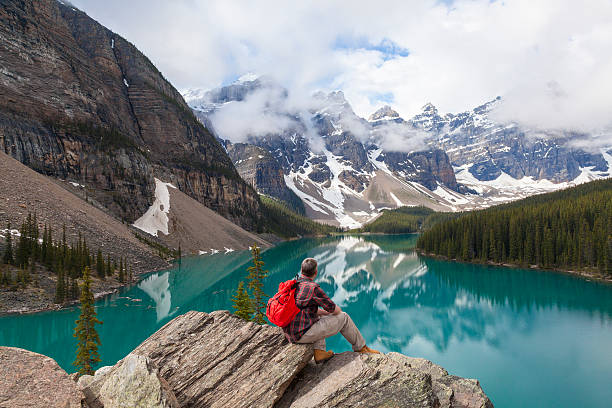Beautiful photo of a man atop rocks looking at Moraine lake Hiking man sitting down with rucksack backpack standing on tree log by Moraine Lake looking at snow covered Rocky Mountain peaks, Banff National Park, Alberta Canada moraine lake stock pictures, royalty-free photos & images