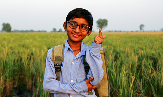Elementary age school student wearing school uniform and carrying school bag thinking something standing portrait near green field during springtime.