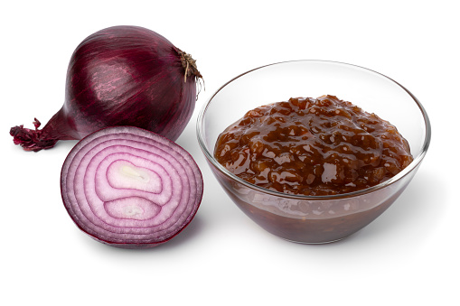 Glass bowl with caramelized red onion chutney and a fresh red onion on the side close up isolated on white background